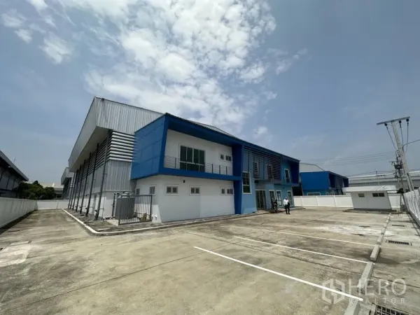 Factory for rent in Bang Sao Thong, Samut Prakan - Modern blue-and-white factory with two-storey office, fenced site and a large concrete parking yard under a bright sky.