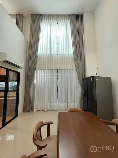 Townhouse for rent in Suan Luang, Bangkok - Double-height window with sheer and blackout curtains beside a sliding door to the kitchen.