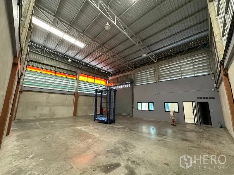 货栈 出租在Khlong Luang, Pathum Thani - 开阔仓库地面，卷闸门、侧边办公室窗户与钢结构屋顶。