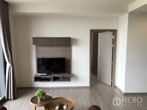 Condominium for rent in Watthana, Bangkok - Modern living area with mounted TV wall unit, light wood floors, and doorway to the bedroom.