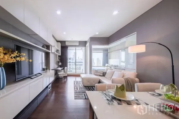 Condominium for rent or sale in Watthana, Bangkok - Open-plan living and dining area with sofa, TV unit and balcony access overlooking the city.