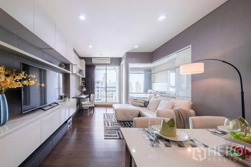 Condominium for rent or sale in Watthana, Bangkok - Open-plan living and dining area with sofa, TV unit and balcony access overlooking the city.