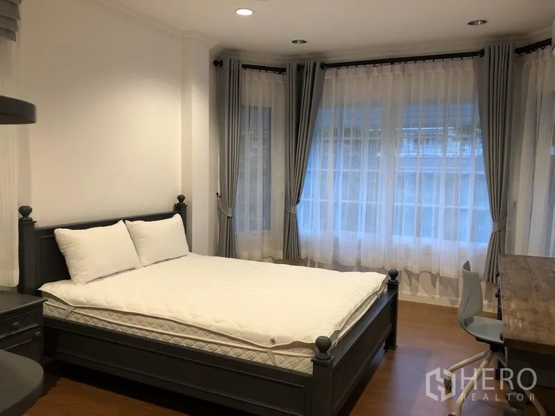Detached House for rent in Mueang Samut Prakan, Samut Prakan - Corner bedroom with dark bed, bay windows dressed in sheer curtains and study desk.
