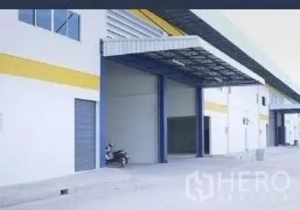 Factory for rent in Mueang Samut Sakhon, Samut Sakhon - Industrial warehouse exterior with wide electric roller shutter, covered loading bay and pedestrian door along a broad driveway.