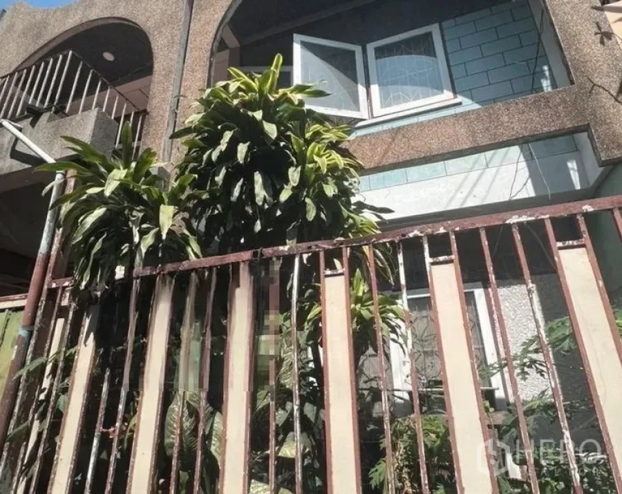Townhouse for sale - Two-storey Ekkamai townhouse facade with arched balcony, open windows, and mature greenery behind a metal gate.