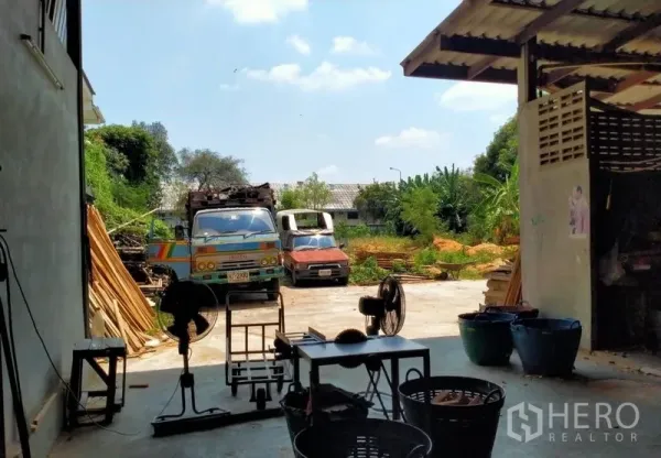 Warehouse for sale in Mueang Nonthaburi, Nonthaburi - Semi-open workshop leading to yard with parked trucks and industrial equipment.