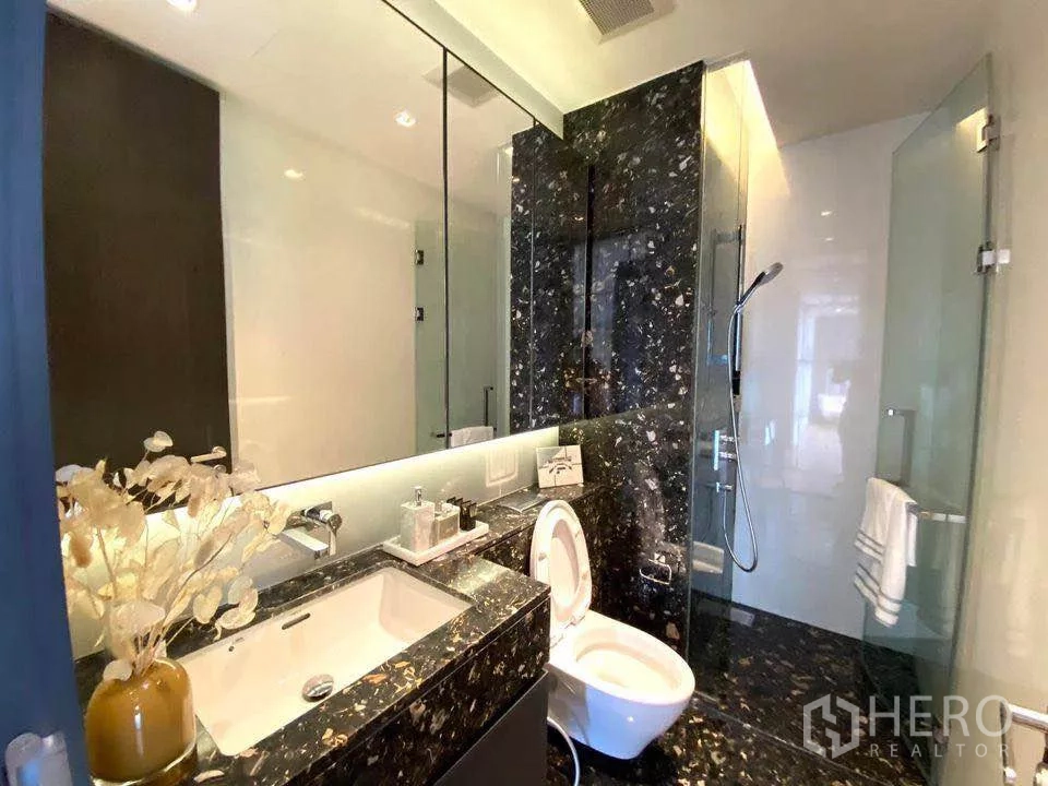 Condominium for rent or sale in Khlong Toei, Bangkok - Bright bathroom featuring glass shower, dark stone accent wall and wide mirror.