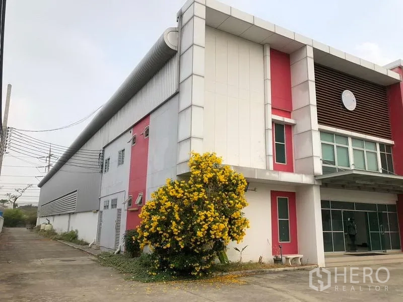Factory for rent in Bang Sao Thong, Samut Prakan - Side view of factory façade with red columns and a flowering shrub by the walkway.