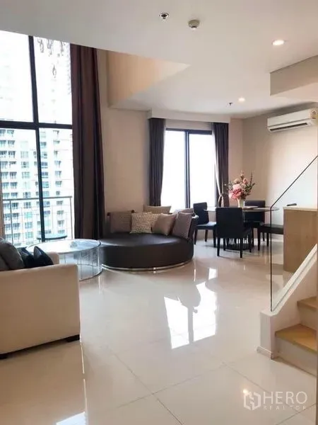 Condominium for rent in Ratchathewi, Bangkok - Bright double-height living room with floor-to-ceiling windows, round sofa, dining set and balcony city view.