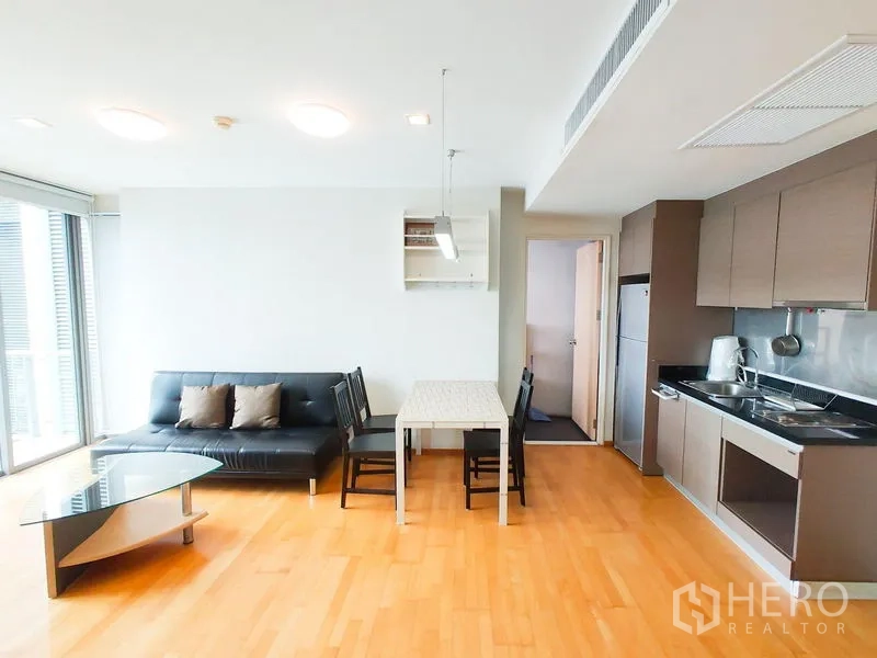 Condominium for rent in Khlong Toei, Bangkok - Open-plan living room with kitchenette, dining table, black sofa and glass coffee table on wood floors.