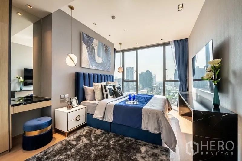 Condominium for rent or sale in Khlong Toei, Bangkok - Master bedroom with blue upholstered headboard, pendant lights, floor‑to‑ceiling windows, and expansive city view.