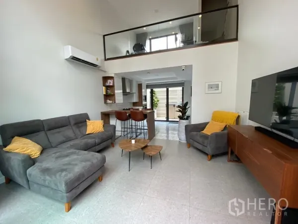 Townhouse for rent in Suan Luang, Bangkok - Open-plan living room with double-height ceiling, grey sectional sofa, TV console and mezzanine.