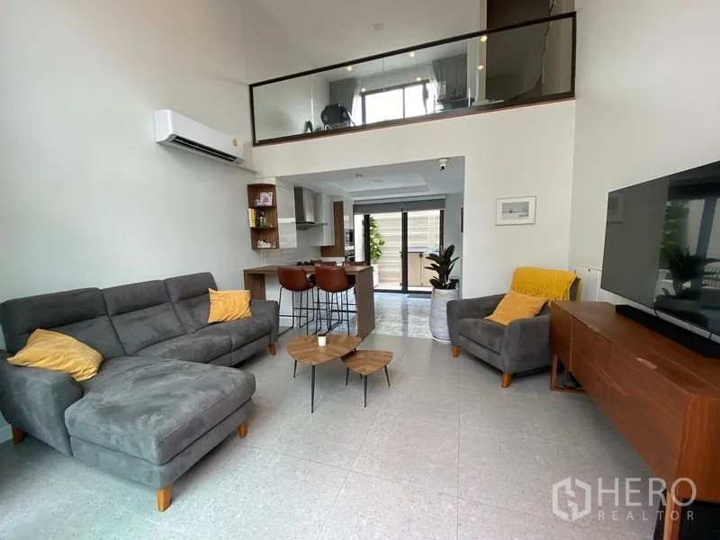 Townhouse for rent in Suan Luang, Bangkok - Open-plan living room with double-height ceiling, grey sectional sofa, TV console and mezzanine.
