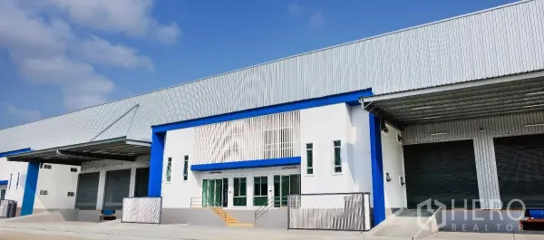Factory for rent in Bang Sao Thong, Samut Prakan - Modern factory exterior with white and blue facade, covered loading bays, and multiple roller shutter doors under a corrugated metal roof.