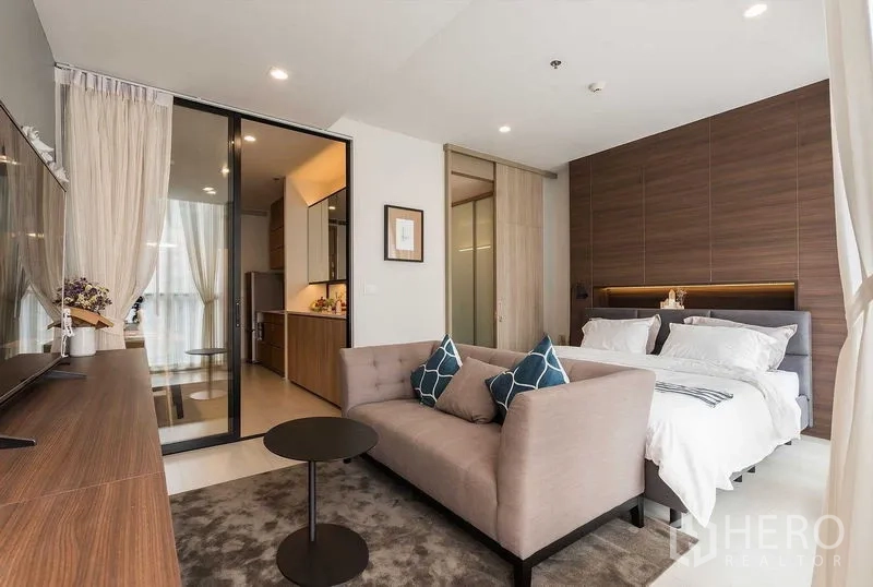 Condominium for rent in Pathum Wan, Bangkok - Open‑plan living and bedroom with sofa, TV console and sliding glass door to the kitchen, framed by sheer curtains.