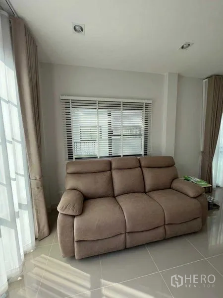 Detached House for rent or sale in Samut Prakan - Sunlit living room corner with reclining sofa, blinds and tile floor.