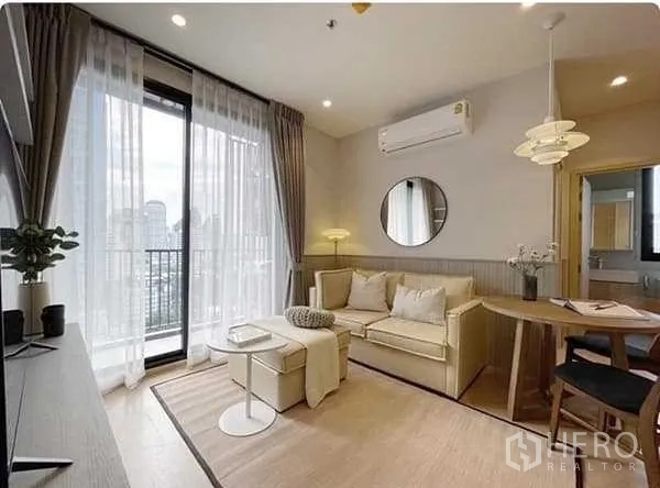 公寓 出租在Watthana, Bangkok - 明亮客厅配米色沙发、圆镜与空调，落地窗外连通阳台可赏城市景观。