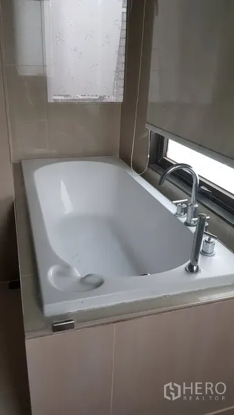 Condominium for rent in Ratchathewi, Bangkok - Bathroom with built-in bathtub positioned beside a window with a roller blind.