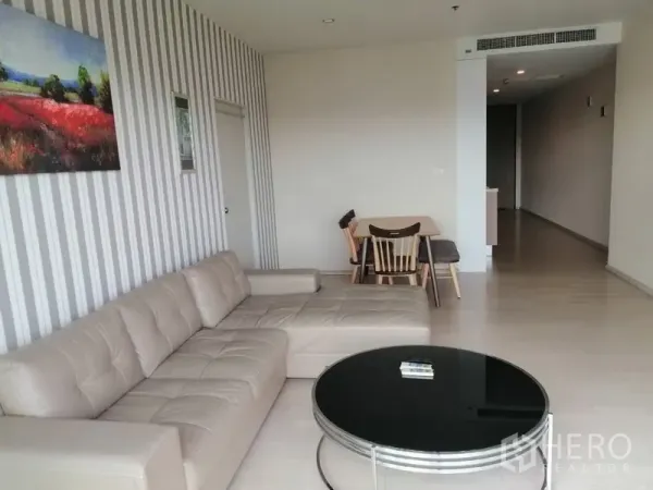 คอนโด ให้เช่าในWatthana, Bangkok - ห้องนั่งเล่นสว่าง พื้นไม้ โซฟาตัวแอลสีครีม โต๊ะกลางทรงกลม และผนังลายทาง พร้อมมุมทานอาหารเล็ก.