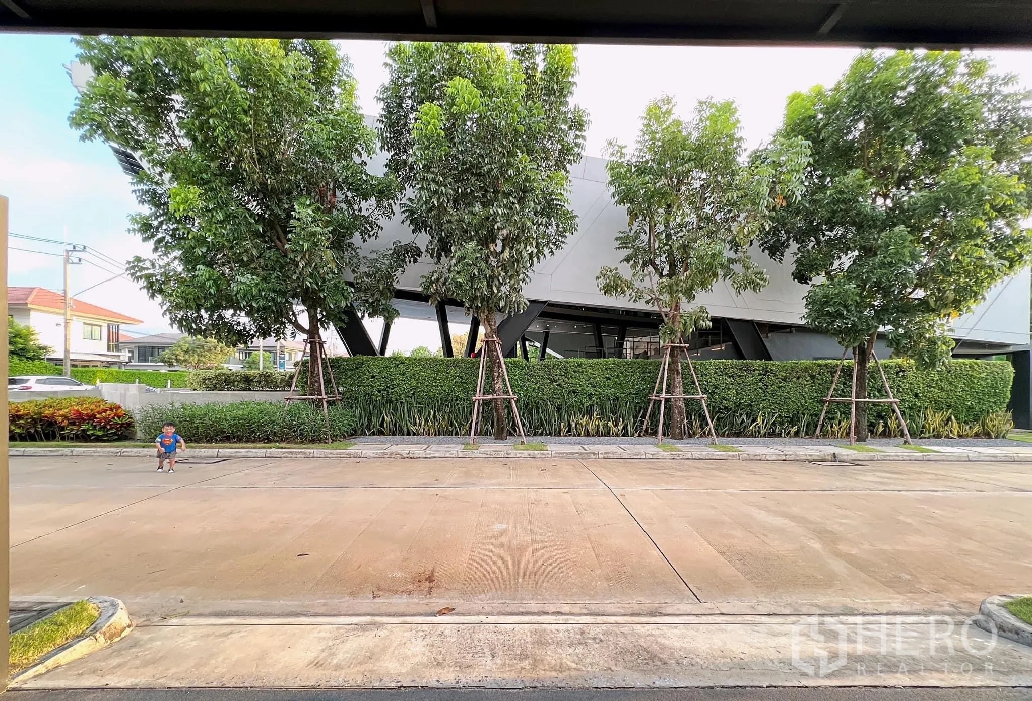 Detached House for rent or sale in Lam Luk Ka, Pathum Thani - View from driveway toward landscaped clubhouse across tree-lined street.