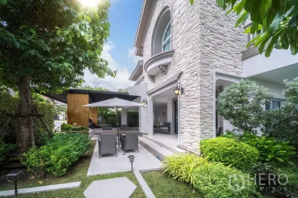 Detached House for rent in Bang Khen, Bangkok - Landscaped garden with patio dining set under umbrella beside the stone-clad exterior.