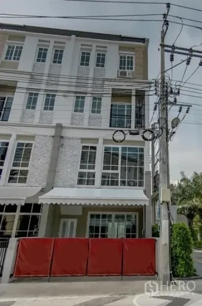 带办公室的家 出租在Bang Na, Bangkok - 街角现代住办合一建筑外观，落地网格窗、白色遮阳篷与红色围栏，临静街。