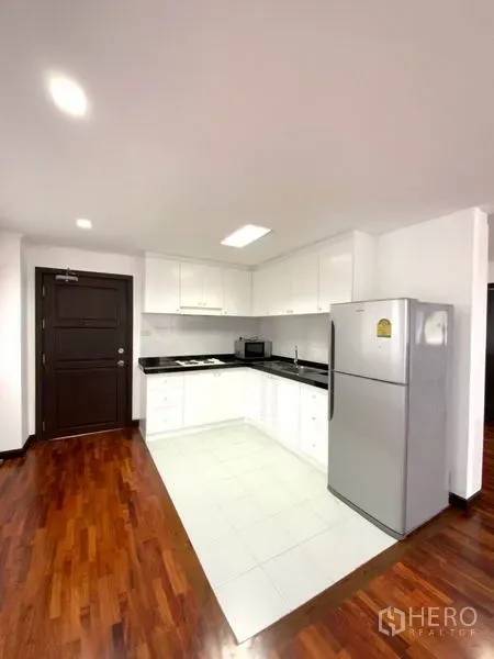 公寓 出租在Watthana, Bangkok - 紧凑白色厨房，黑色台面、吊柜、灶台与大冰箱，靠近入户门。