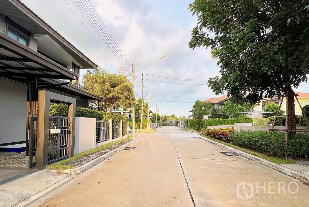 Detached House for rent or sale in Lam Luk Ka, Pathum Thani - Quiet residential street inside a gated estate with trees and sidewalks.