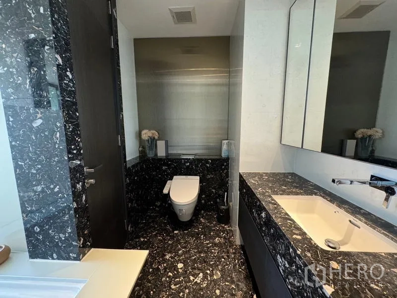 Condominium for rent in Khlong Toei, Bangkok - Contemporary bathroom with black marble surfaces, wall-hung toilet and wide vanity.