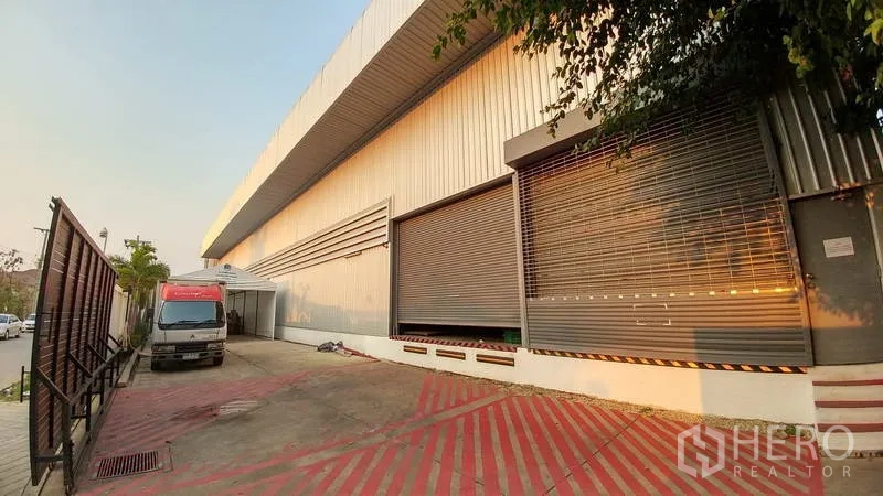 Warehouse for rent or sale in Nonthaburi - Exterior loading zone with roller shutters and truck access beside the warehouse.