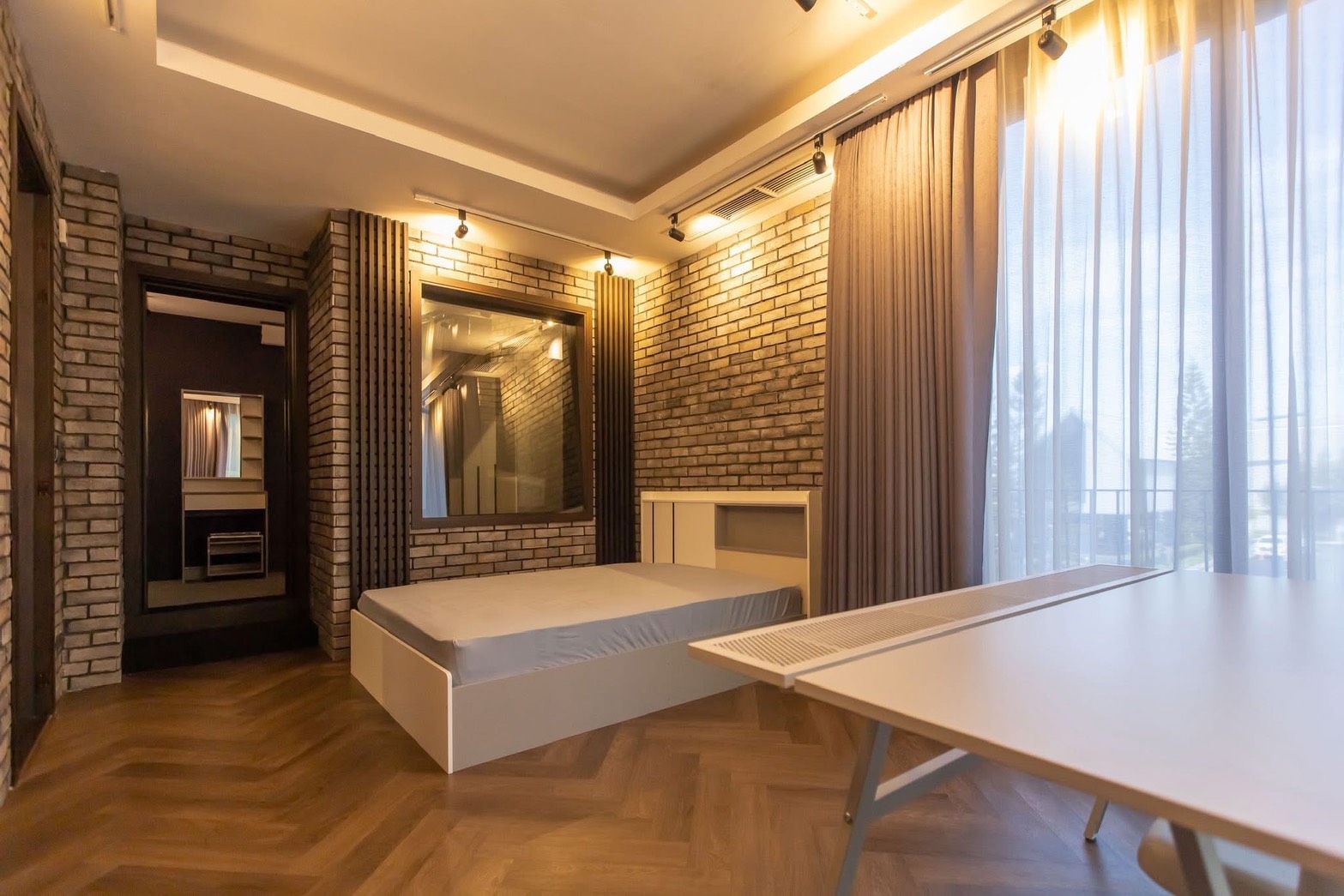 Detached House for rent in Lat Phrao, Bangkok - Warm bedroom with brick feature wall, platform bed, herringbone wood floor, and sheer curtains.