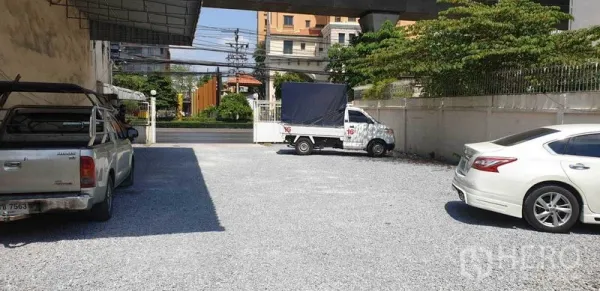 Shophouse for rent in Phra Khanong, Bangkok - Spacious gravel parking yard with cars and a pickup truck inside a gated property facing the main road.