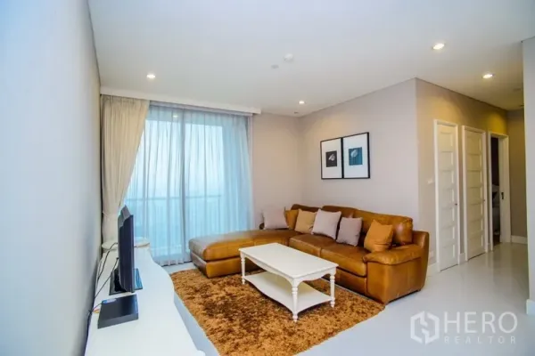 Condominium for rent in Khlong Toei, Bangkok - Bright living room with a leather sectional sofa, white coffee table, TV console and floor-to-ceiling window to a balcony view.
