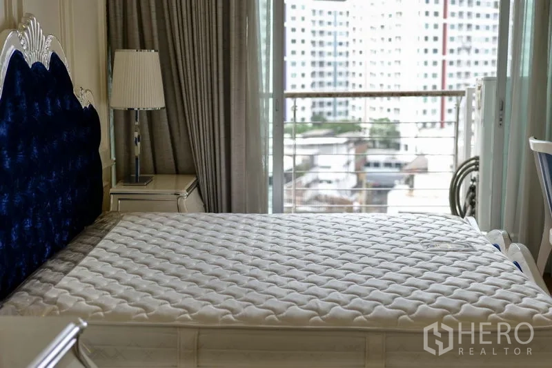 Condominium for rent in Khlong Toei, Bangkok - Bedroom with tufted blue headboard and balcony doors opening to a city view.