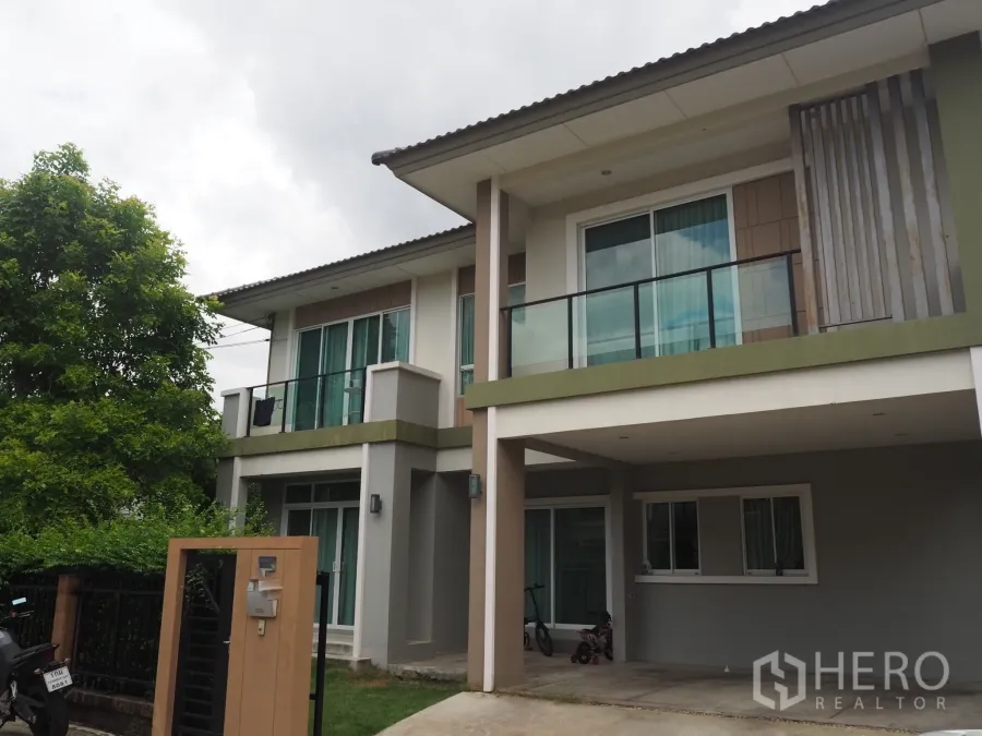 独立式住宅 出租在Pathum Thani - 两层独栋住宅外观，带阳台、遮雨车位、入户门与绿树环绕。