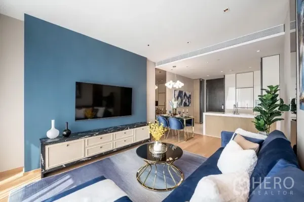 Condominium for rent or sale in Khlong Toei, Bangkok - Open‑plan lounge and dining area with blue accent wall, marble TV console, round coffee table, and view toward the kitchen.
