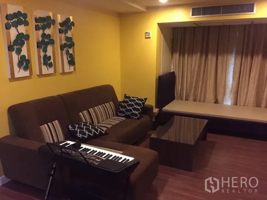 Condominium for rent in Watthana, Bangkok - Cozy living room with brown sofa, wood floors, yellow walls, wall art, coffee table and a curtained window seat.