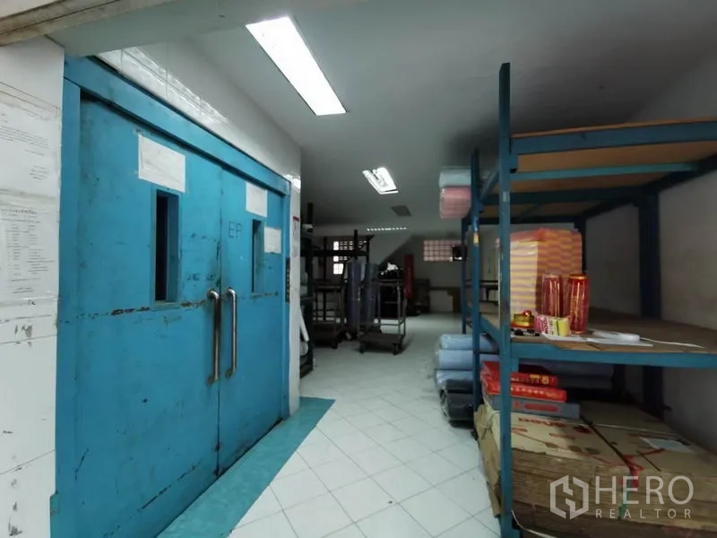 办公空间 出租在Khlong San, Bangkok - 仓储区，蓝色双开工业门与金属货架，荧光灯照明。