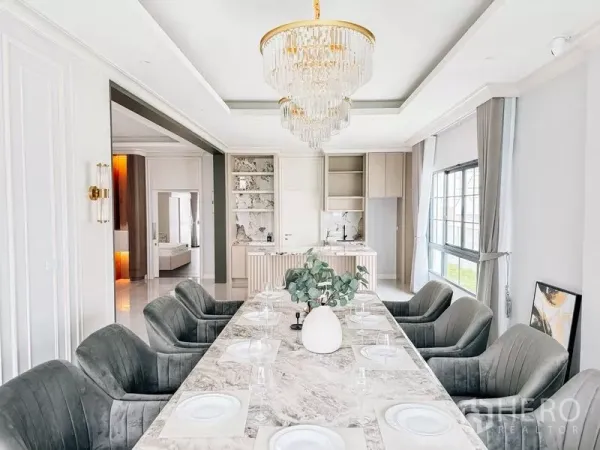 Detached House for rent or sale in Bang Phli, Samut Prakan - Elegant dining room with long marble table, upholstered chairs and crystal chandelier beside a pantry kitchen.