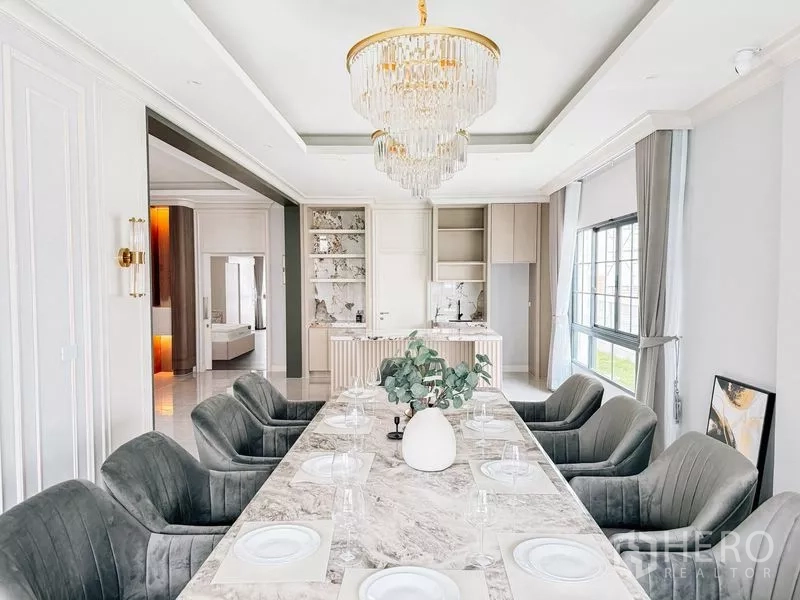 Detached House for rent or sale in Bang Phli, Samut Prakan - Elegant dining room with long marble table, upholstered chairs and crystal chandelier beside a pantry kitchen.