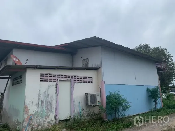 Warehouse for rent in Lam Luk Ka, Pathum Thani - Warehouse exterior with corrugated metal siding, wall-mounted air conditioner, satellite dish, and weathered paint.