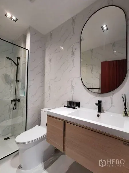 Townhouse for rent or sale in Yan Nawa, Bangkok - Modern bathroom with glass shower, marble-look walls, vanity with oval mirror and black fixtures.