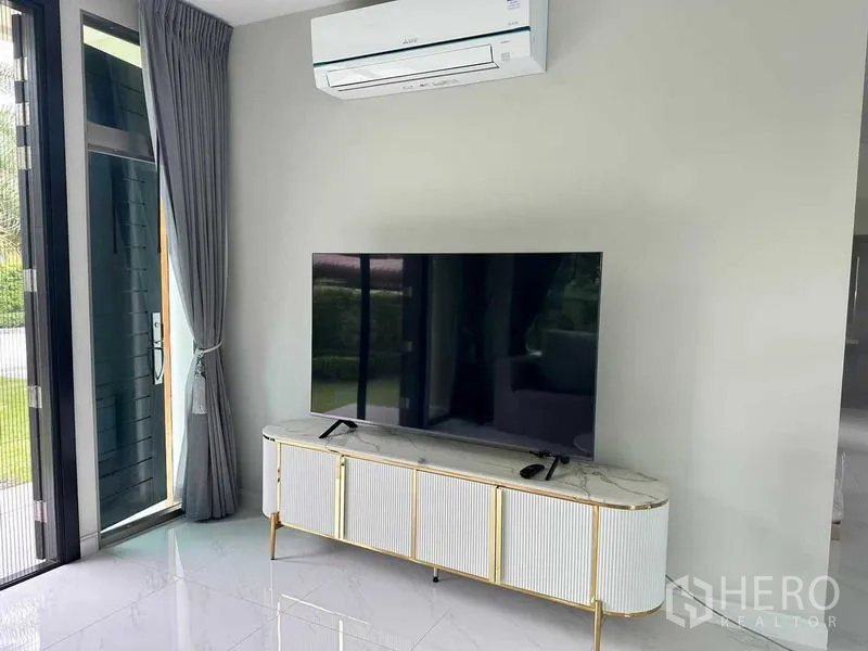 Detached House for rent in Lat Krabang, Bangkok - Living area with large TV on a marble-topped console, sliding glass door, curtains and air conditioner.