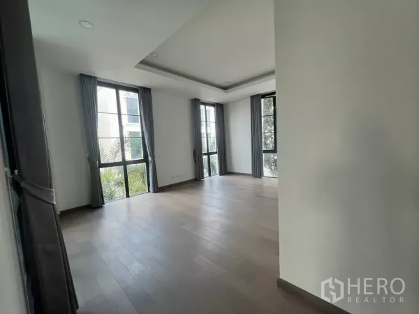 Detached House for rent in Lat Phrao, Bangkok - Large corner bedroom with wood flooring and tall windows with grey curtains overlooking greenery.