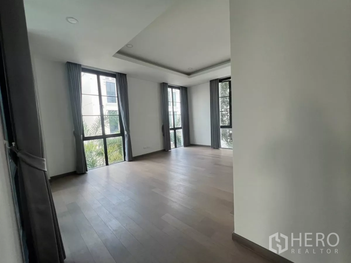 Detached House for rent in Lat Phrao, Bangkok - Large corner bedroom with wood flooring and tall windows with grey curtains overlooking greenery.