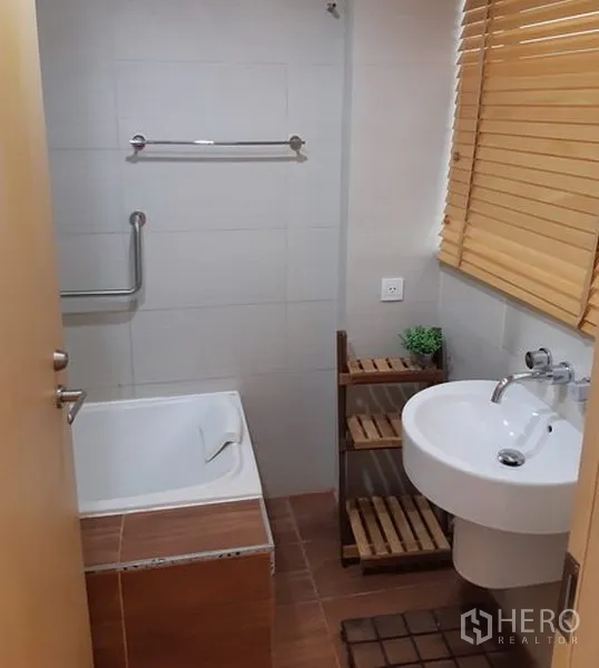 Condominium for rent in Khlong Toei, Bangkok - Bathroom with bathtub, pedestal sink, wooden shelving and window blinds.