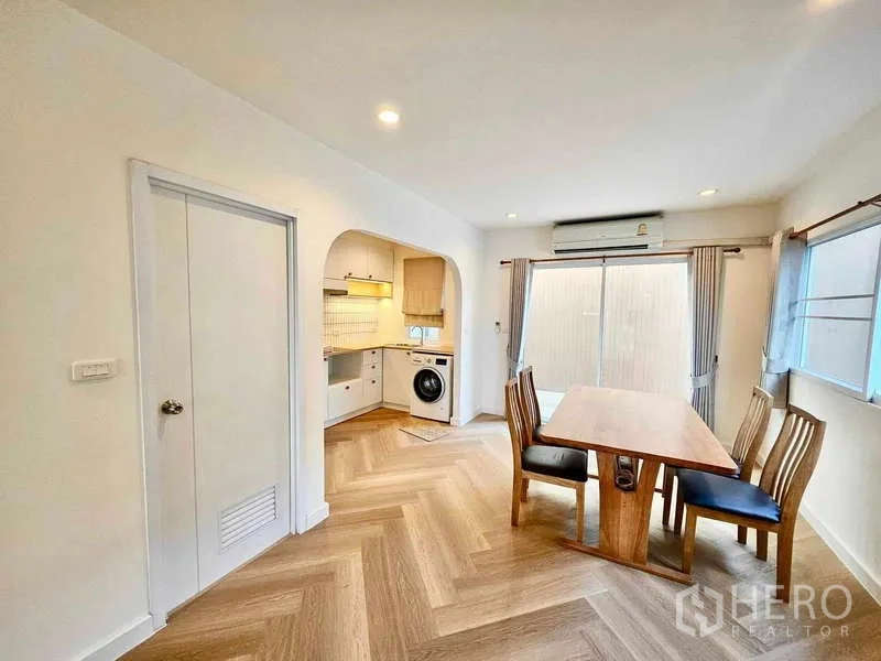 Townhouse for rent in Suan Luang, Bangkok - Open dining area with a wooden table, windows, and a sliding door next to an arched entry leading to the fitted kitchen.