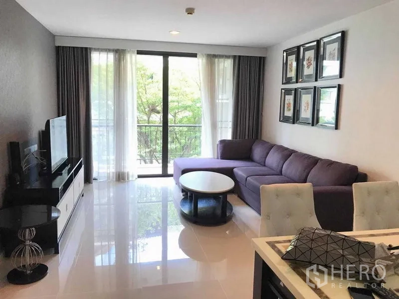 Condominium for rent in Khlong Toei, Bangkok - Open living room with L-shaped sofa, TV console, and sliding doors to a green-facing balcony.
