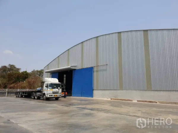 货栈 出租在Chaloem Phra Kiat, Saraburi - 大型工业仓库侧视图，蓝色入口与宽大铺装回旋区，适合拖车调头，沙拉武里。