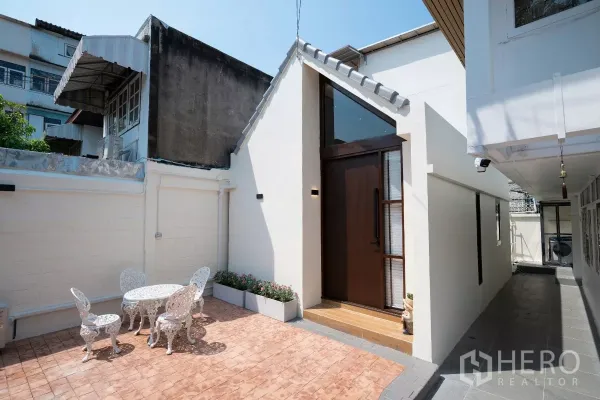 Detached House for rent in Phra Khanong, Bangkok - White gable-front detached house with tall wooden door and sunny brick-paved courtyard with patio set.